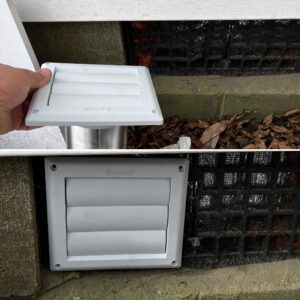 A person is installing a white vent cover onto a rectangular wall opening. The opening is surrounded by a mixture of concrete and a wire mesh, with dried leaves visible inside.