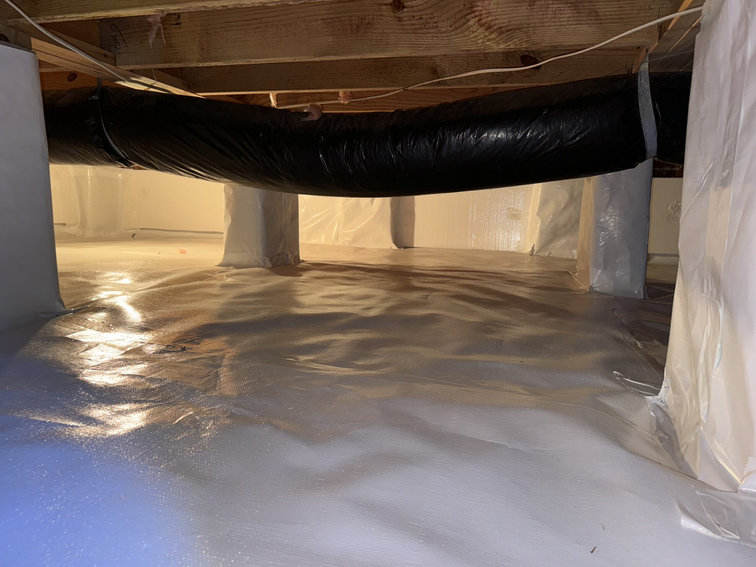 A clean and well-lit crawl space with a plastic vapor barrier covering the floor and parts of the support columns, and an insulated duct running across the ceiling.