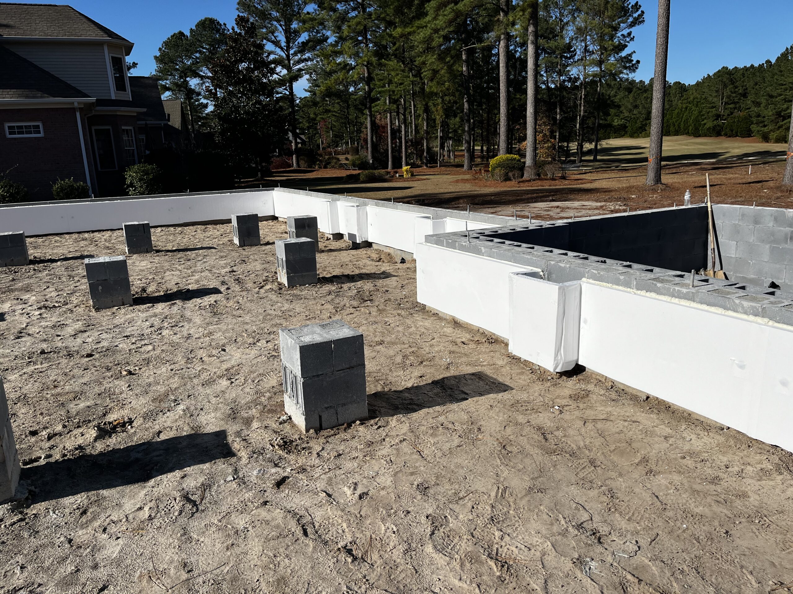 A building's concrete foundation under construction surrounded by trees. Rectangular concrete blocks are placed on the ground, and partial walls are in progress.