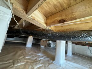 A crawl space with insulated ductwork, plastic-wrapped support pillars, and a dehumidifier visible in the background. The wooden beams above support the floor above.