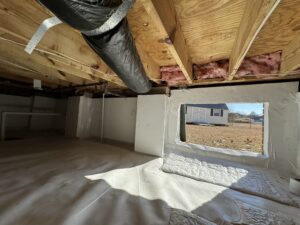 A view of a well-lit crawl space with wooden beams, insulation, and a large open window. A black duct runs along the ceiling, and a barn is visible outside.