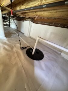 A crawl space with a white plastic moisture barrier installed on the ground and walls, featuring pipes, wooden beams overhead, and a black circular base with a pipe in it.