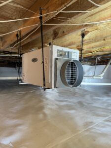 A dehumidifier is installed in a basement with exposed wooden beams and a white plastic-lined floor.