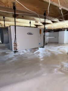 A white dehumidifier unit is installed in a crawl space with wooden beams above and plastic lining the floor and walls. Cables and cords are visible.