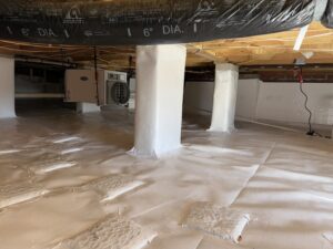 A clean, well-lit basement with white plastic-wrapped support columns, an air conditioning unit, and insulated flooring.