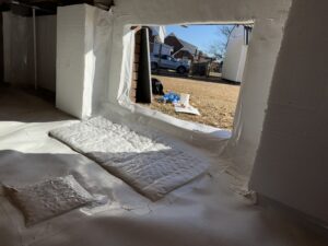 A basement area with white insulation and a cut-out window revealing an outdoor view, including a house, lawn, and some rolled-up insulation materials on the ground.