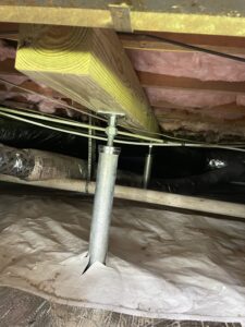 A view of a house crawl space showing an adjustable metal support post secured to a wooden beam, with pink insulation and ductwork visible in the background.