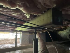 Crawl space with exposed insulation, wooden beams, and various pipes and supports. Dim lighting reveals dirt floor and plastic sheeting.