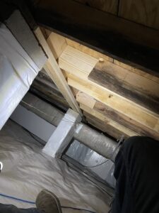 View of an attic with exposed wooden beams and insulation. A wrapped duct runs through the space. The bottom edge of a person's shoes is visible in the lower part of the image.