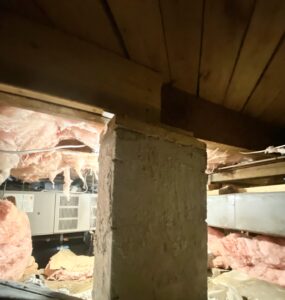 A concrete pillar supports wooden beams in a basement with exposed insulation and HVAC units in view.