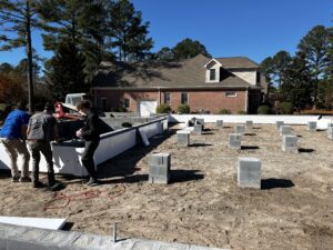 Workers at a residential construction site are preparing the foundation, with concrete blocks in place and tools on the ground. A house and trees are visible in the background under a clear sky.