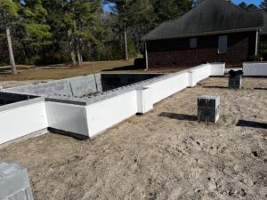 A brick foundation under construction with white insulation panels is shown alongside a house in the background.