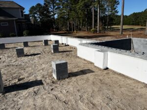 Concrete foundation with block columns on a sandy construction site, bordered by trees and a residential building.