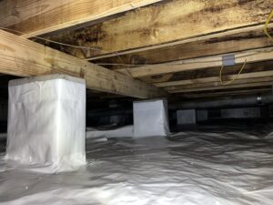 A crawl space with wooden beams, concrete supports wrapped in white material, and a plastic-covered floor.