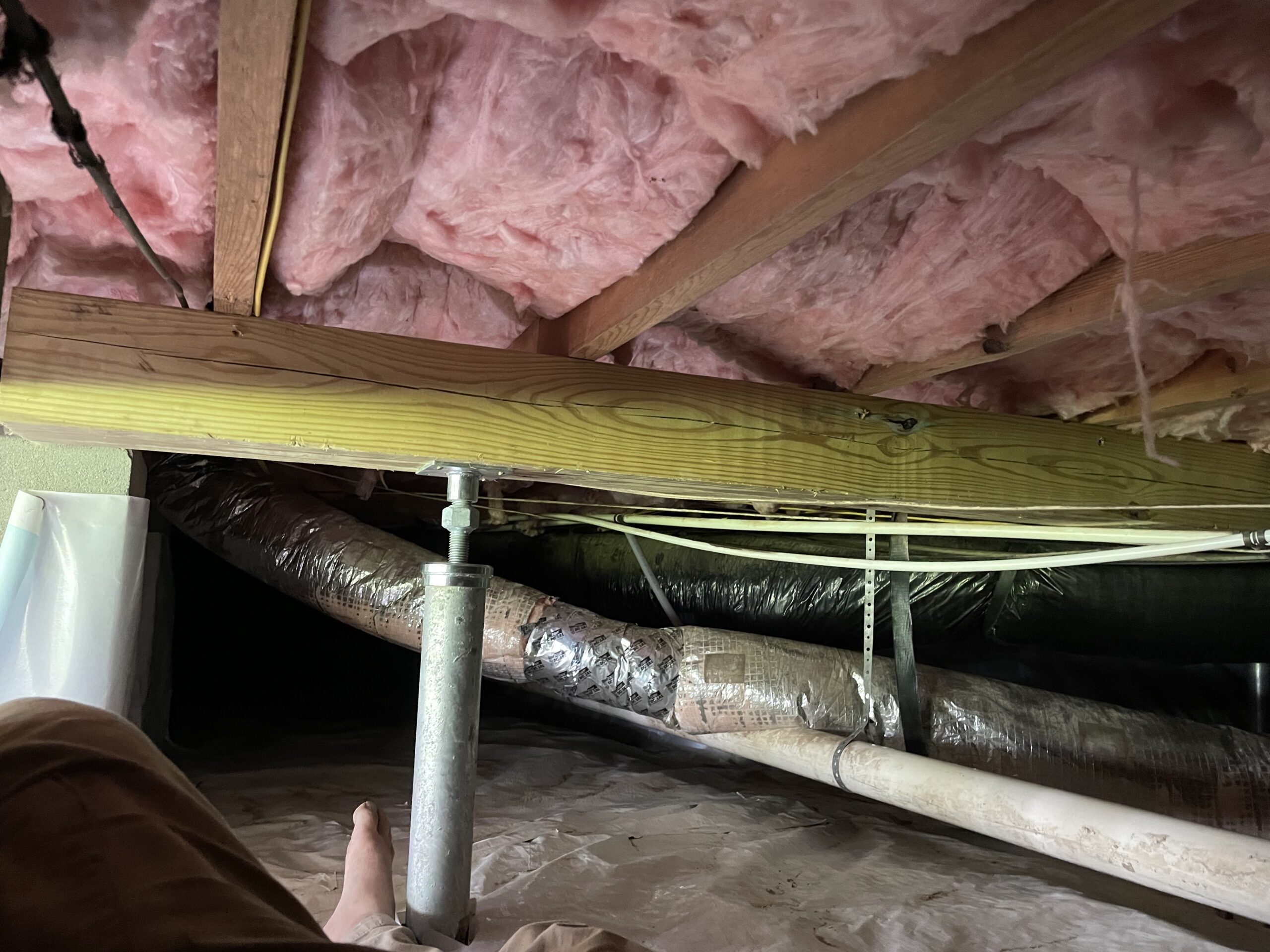 A view of an insulated ceiling in a crawl space with ducts, pipes, and a worker's foot visible in the foreground.