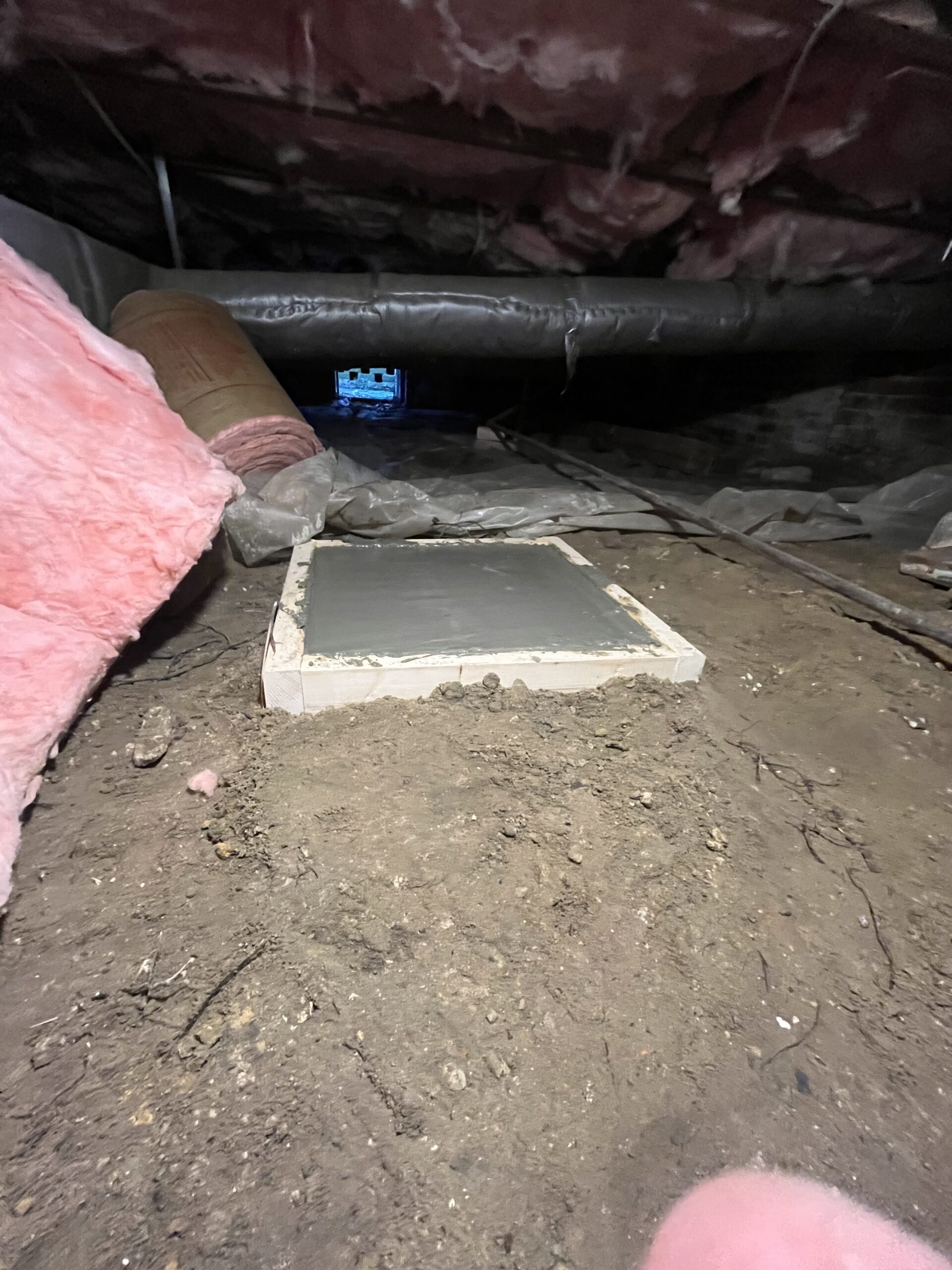 A freshly poured concrete slab in an unfinished attic space surrounded by insulation materials and exposed dirt.